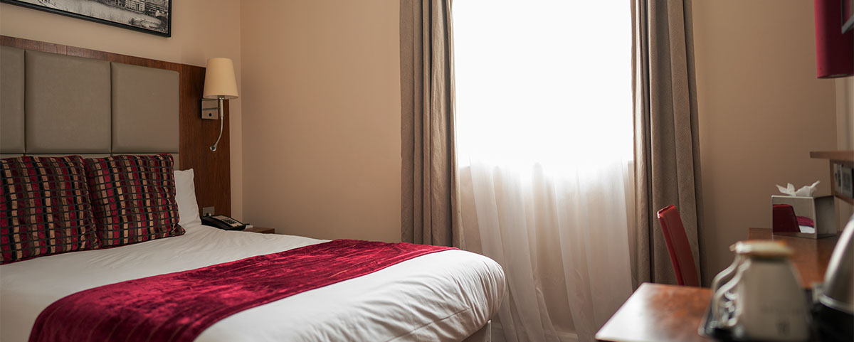 A large bed with a white duvet, red patterned cushions and red bed throw with a city view in the window.